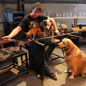 Artist works on glass piece with 3 dogs surrounding him