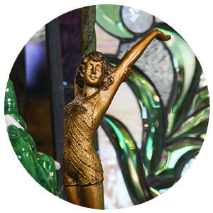 Circle crop of an antique gold painted sculpture of a girl with flowing hair, stretching her arms in a swimsuit.