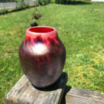 Outdoor shot of a red and black glass vase by Joe Mattson. In the background is a grassy green yard and a chain link fence