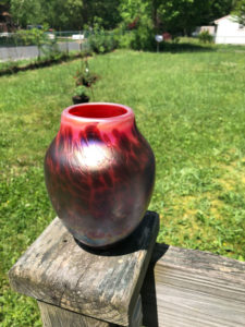 Outdoor shot of a red and black glass vase by Joe Mattson. In the background is a grassy green yard and a chain link fence