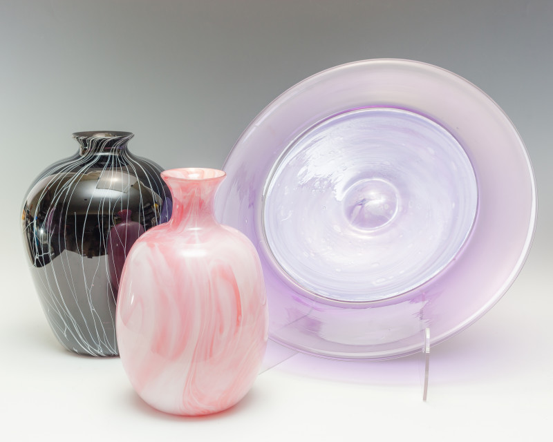 One black glass vase with white detailing, one marbled pink glass vase, and one light purple glass plate on display stand.