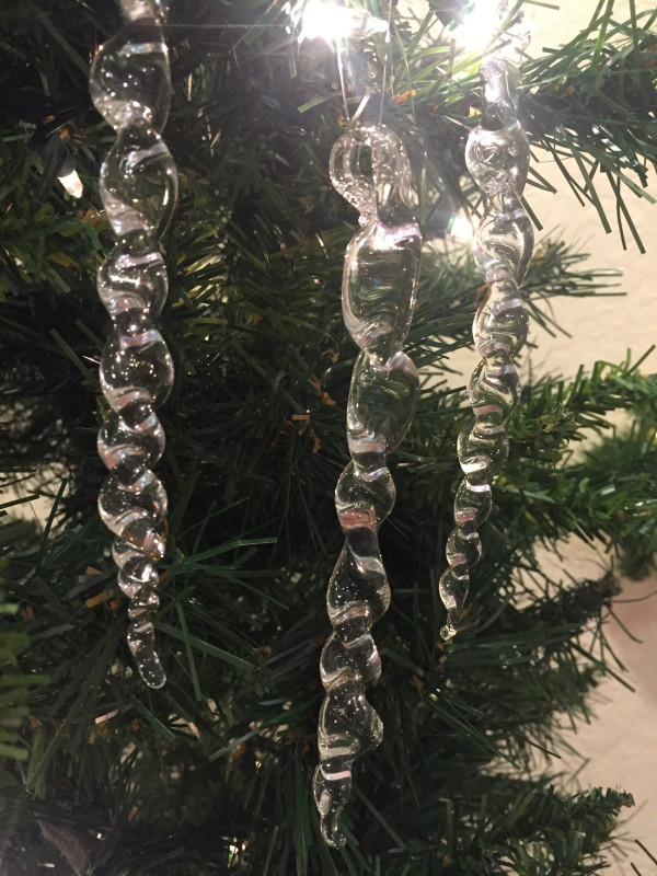 Light reflecting off of three glass icicles, hanging from a pine tree in front of a white wall.