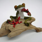 A sculpture of a stylized frog holding two red lizards. They sit on a tan piece of driftwood. Created by Sandy Lehman.