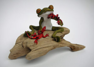 A sculpture of a stylized frog holding two red lizards. They sit on a tan piece of driftwood. Created by Sandy Lehman.