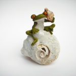 A sculpture of a stylized frog listening to a tiny conch shell. The frog sits on a white spiral shell. Created by Sandy Lehman.