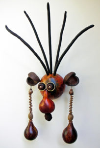 Carved gourd face by Robert Lehman with tall black pieces of hair, large ears with larger dangling earrings, and a hanging nose.