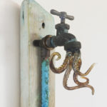 An outdoor faucet fixed to a piece of wood, flailing glass octopus legs come out of the faucet. By Scott Manns