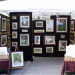 A collection of Ramona Maziarz's paintings in a booth setting. Each painting is in a deep colored frame with white matting.