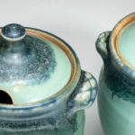 Closeup of two matching teal ceramic vessels, each with dark teal accents around the rim and lid. Created by Amy Peseller.