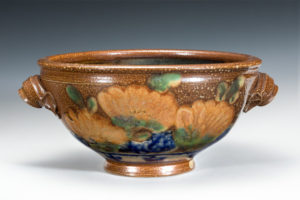 Large brown ceramic bowl with handles. Flowers painted into the glaze around the base of the bowl. Created by Terry Plasket.
