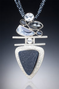 A silver pendant by Willie Trejbal with several faceted black and greystones stacked over two bars and a black triangular stone at the base.