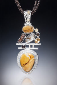 A silver pendant by Willie Trejbal with several faceted orange and brown stones stacked over two bars and an orange teardop shaped stone at the base.