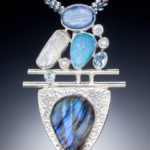 A silver pendant by Willie Trejbal with several smooth blue and white stones stacked over two bars and a tear drop shape stone at the base.