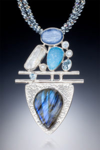 A silver pendant by Willie Trejbal with several smooth blue and white stones stacked over two bars and a tear drop shape stone at the base.