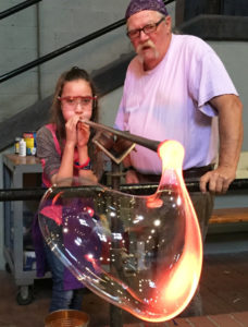 Glass artist Joe Mattson instructing a young girl how to blow a large glass bubble with a blowpipe.