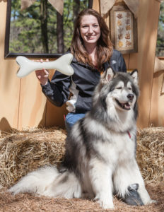 Woman sitting on straw, holding a large glass bone with her dog during PAWS for art.