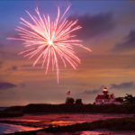 Photograph taken by Dennis Abriola of a bright pink firework exploding over the beach at sunset, with an american flag and lighthouse in the distance.