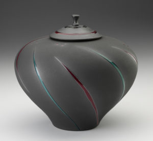 A large lidded grey jar with a wide top, tapering like a funnel to a smaller base. Single curves of alternating green and red go around the piece from bottom to top. Created by Michael Cho