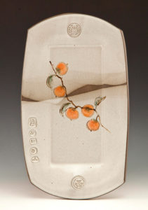 A long white ceramic piece depicting fruits on a branch along its center. Created by Pamela Cummings
