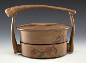 A ceramic piece with a large circle lid, a fruit motif, and a large handle. Created by Pamela Cummings