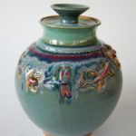 A deep turquoise color pot with a line of various creatures around the body, such as a dragonfly, lizard, and frog. Created by Marsha Dowshen