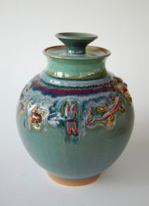 A deep turquoise color pot with a line of various creatures around the body, such as a dragonfly, lizard, and frog. Created by Marsha Dowshen