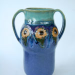 A tall vase with two handles on either side, the top is a deep turquoise, changing to a navy blue as it goes down. Around the center are three decorative sunflowers. Created by Marsha Dowshen