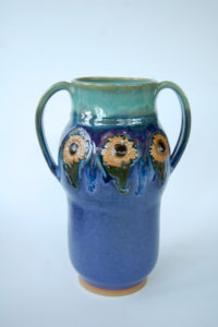 A tall vase with two handles on either side, the top is a deep turquoise, changing to a navy blue as it goes down. Around the center are three decorative sunflowers. Created by Marsha Dowshen