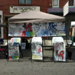 A collection of artwork by Daniel Duffy in a booth setting, under a tent with prints arranged in three metal display pieces.