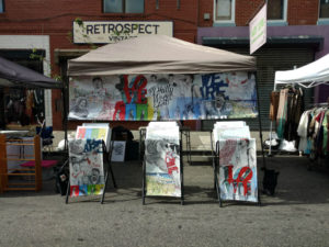 A collection of artwork by Daniel Duffy in a booth setting, under a tent with prints arranged in three metal display pieces.
