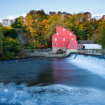 Photograph taken by James Evangelista of a red water mill house across from a large body of swirling water with a small waterfall.
