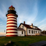 Wispy white clouds in a blue sky create a contrast against the warm colors of the large red and white striped lighthouse with a home attached to it. Photograph taken by James Evangelista