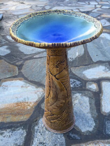 A tan bird bath with an azure basin, highlighted by sunlight. Throughout, there are etchings of leaves of various types. Created by Stephen Fabrico.