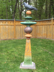 Tall garden sculpture of a raven resting on an earth toned pillar etched with botanical elements. Created by Stephen Fabrico.
