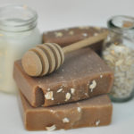 A honey stirrer laying on two bars of soap with two glass bottles in the background. From Bay Berry Bliss LLC