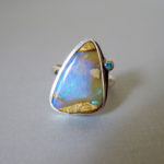 Silver ring by Yanina Siani holding a small faceted gemstone and a larger smooth triangle blue opalescent stone