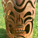 A cylindrical metal garden sculpture with a tiki mask pattern cut into the sides. Created by Sandra Webberking.