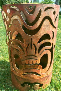 A cylindrical metal garden sculpture with a tiki mask pattern cut into the sides. Created by Sandra Webberking.