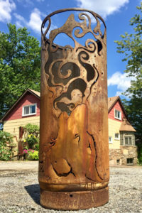 A cylindrical metal garden sculpture with a wolf howling a smoke pattern cut into the sides. Created by Sandra Webberking.