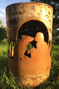 A cylindrical metal garden sculpture with a scene of a girl and her dog in the woods cut into the sides. Created by Sandra Webberking.