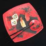Pieces of sushi and a set of chopsticks sit on a rich red sushi plate with small bowl for soy sauce or wasabi. Created by Susan Wechsler