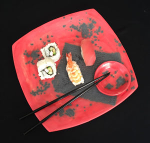 Pieces of sushi and a set of chopsticks sit on a rich red sushi plate with small bowl for soy sauce or wasabi. Created by Susan Wechsler