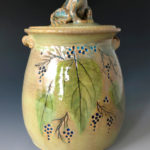 A yellow green ceramic jar with a sculpted toad top by Deborah Williams depicting hanging berries and large green leaves
