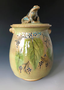 A yellow green ceramic jar with a sculpted toad top by Deborah Williams depicting hanging berries and large green leaves