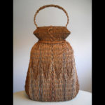 A tall basket with a handle around the lip. The palms are woven to create a zigzag pattern across the middle. Created by Samuel Yao.
