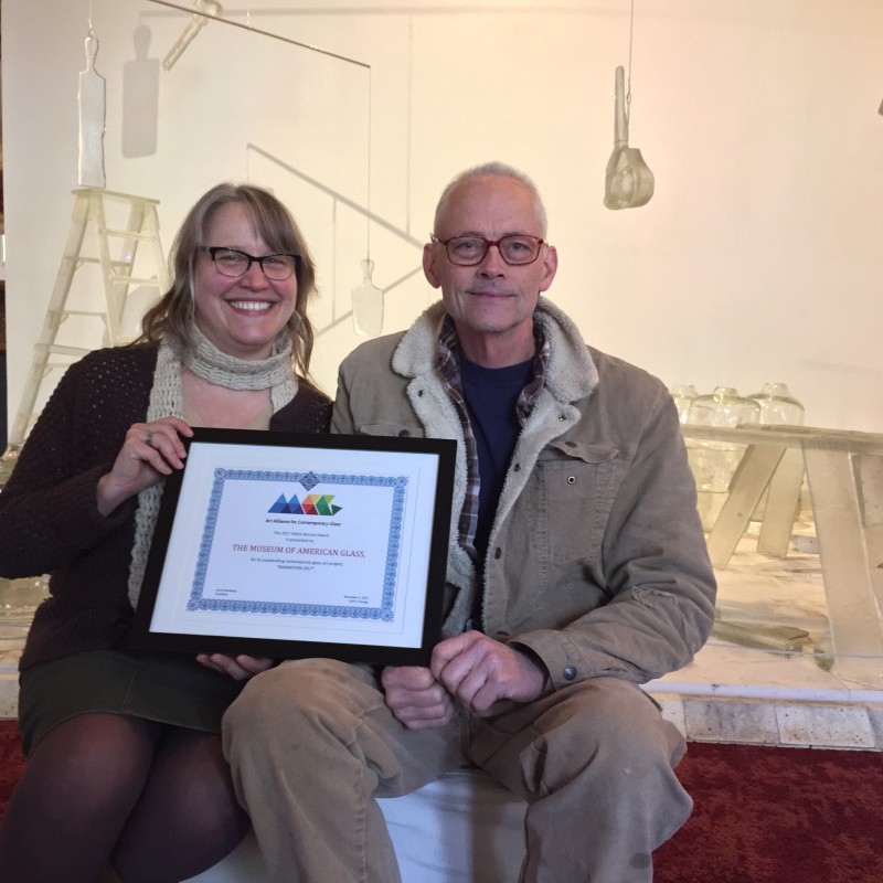 Kristin Qualls sits with Hank Adams, holding a certificate from AACG to the Museum of American Glass