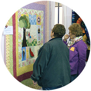 Two women observing a hanging quilt of a landscape scene,