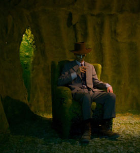 Fellow Bryan McGovern Wilson in a suit and hat, sitting in a green chair, surrounded by a green cavern.