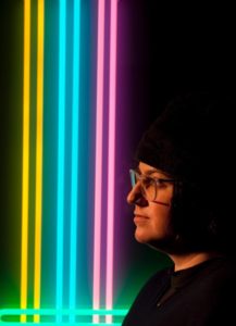 Headshot of Jen Elek in front of three sets of neon lights in yellow, cyan, and magenta. Symbiotic Spheres exhibiting artist.