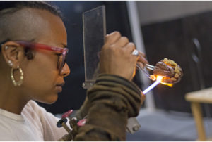Closeup of artist Kimberly Thomas working on a glass jaw behind a small layer of glass, Symbiotic Spheres exhibiting artist.
